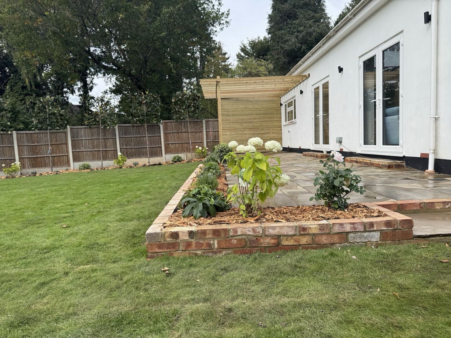 A sandstone patio with a brick-edged raised bed planted up, and a slatted timber screen giving the seating area some privacy from the neighbouring plot.