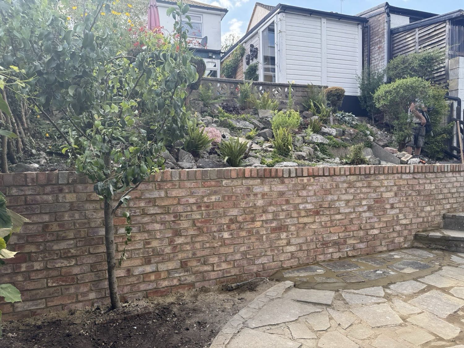 A reclaimed-brick retaining wall holding a raised rockery bed above a crazy-paved path — the footing and the drainage behind it are what keep the face plumb.